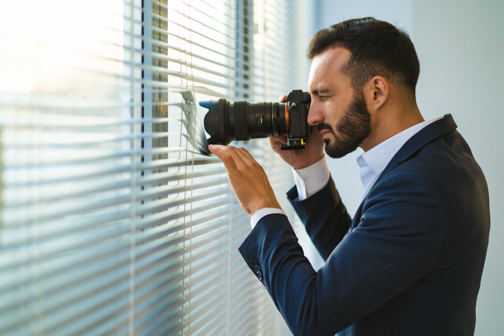 A man with a camera peers through the blinds