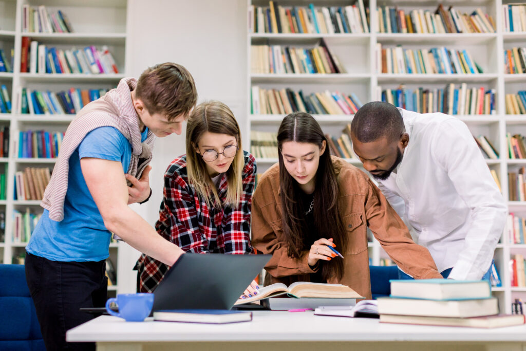 Front view of four concentrated multiethnic young people, college students preparing their homework in a library room, looking for needed information in book. Laptop and books on the table.
